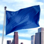 Blue flag made of durable nylon flying on a pole above an urban skyline, Solid Colored Flags - Premier Nylon.