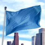 Blue solid colored flag made of premier nylon waving on a flagpole against a city skyline and bright sky, Solid Colored Flags - Premier Nylon.