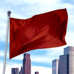 Red flag made of durable nylon fabric flying on a pole against a city skyline, showcasing Solid Colored Flags - Premier Nylon.