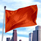 Bright orange flag made of durable nylon waving on a silver pole against a city skyline, Solid Colored Flags - Premier Nylon.
