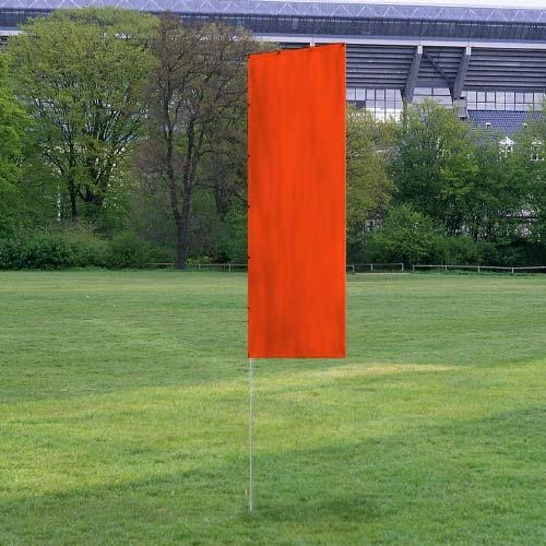 Bright orange solid tall advertising flags standing upright on green grass field with trees and a stadium structure in the background
