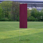 Solid Tall Advertising Flags in solid burgundy color standing upright on a grassy field with trees and a stadium structure in the background.