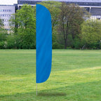 A bright blue Solid Color Flutter Flags standing upright on a grassy field with trees and a building in the background.