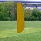 Solid Color Feather Flags in yellow standing upright on a grassy field with trees and a stadium structure in the background.