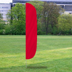 Solid Color Flutter Flags in vibrant red standing upright on a grassy field with trees and buildings in the background.
